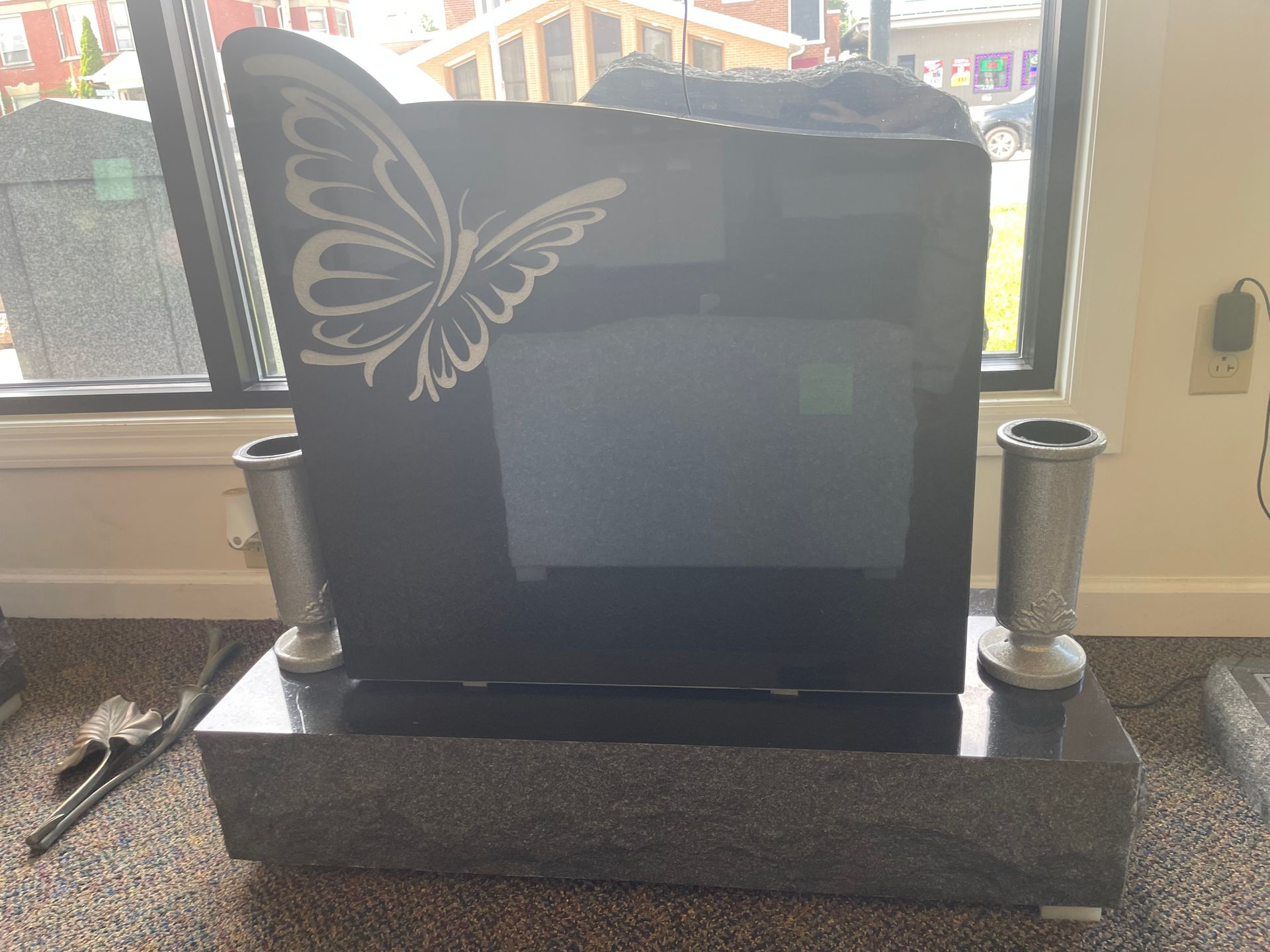 Black granite headstone with a butterfly design and two vases, displayed indoors by a window.