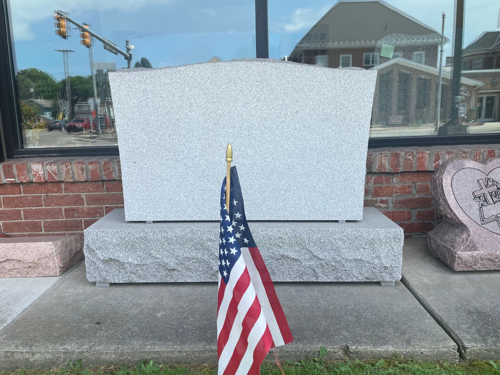 Granite headstone with an American flag in front.  The stone is set on a base and the window behind it reflects a street scene.