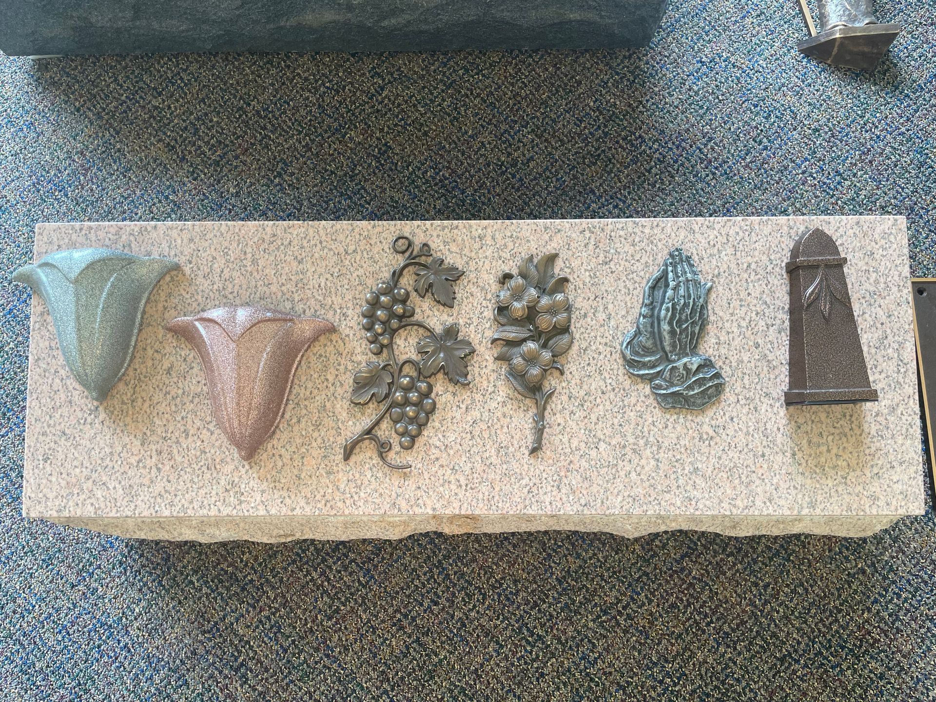 A granite headstone with five decorative carvings: two flowers, grapes, praying hands, and a pillar.