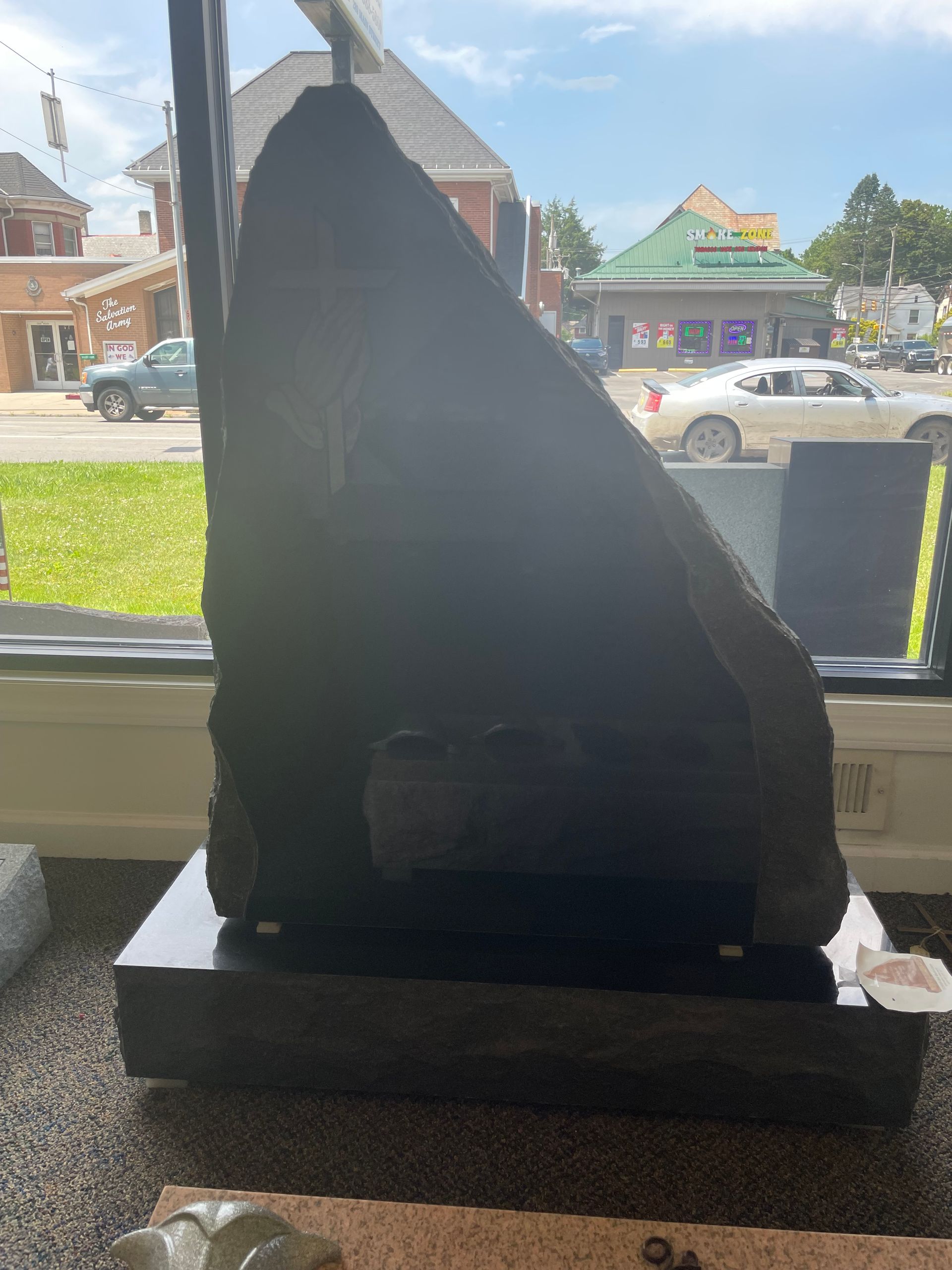 A large black granite monument with a rough, angled top. It sits on a black base, displayed in a shop window with a street view in the background.