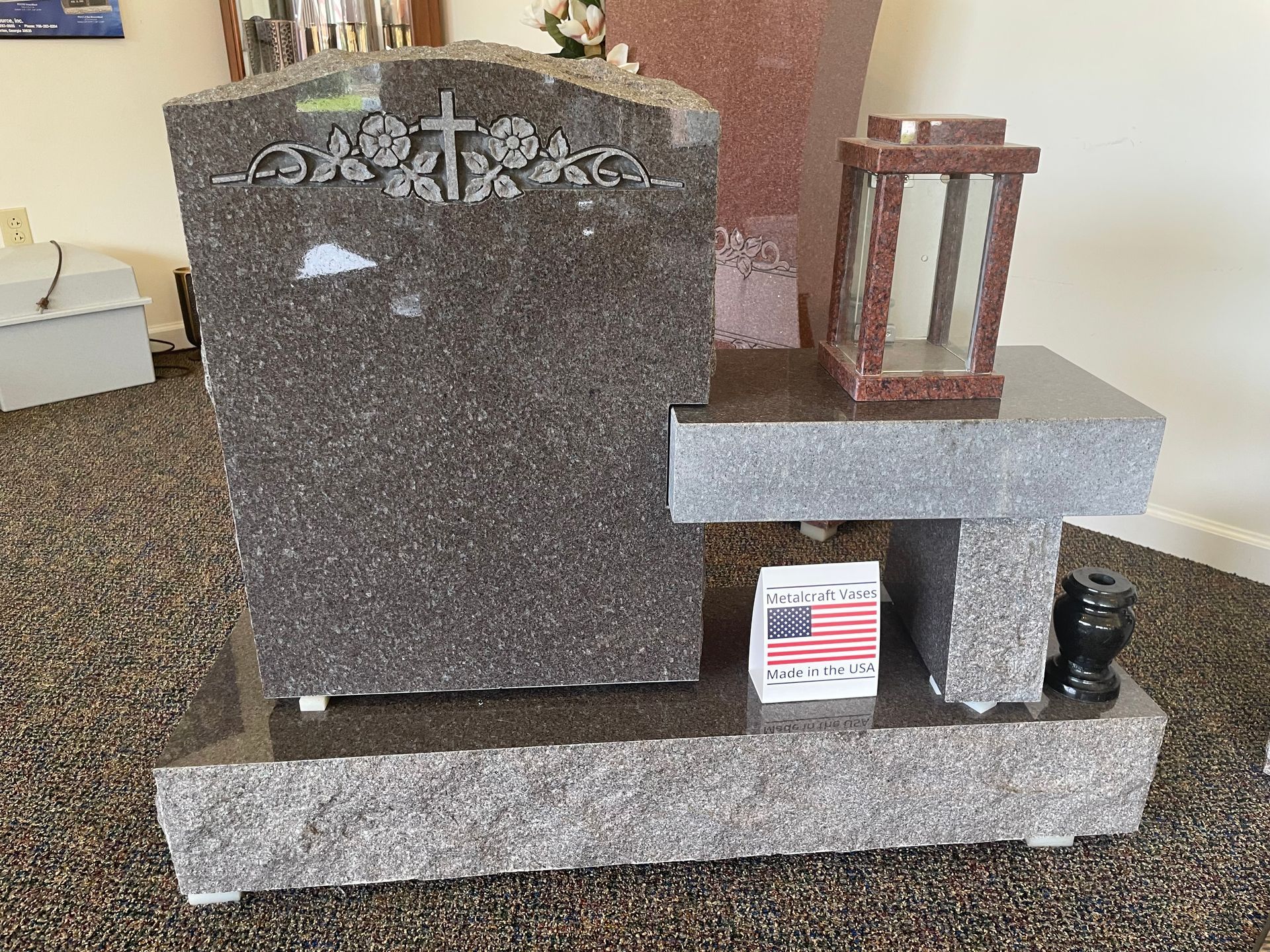 Granite headstone with a cross, flowers, and a lantern, alongside a vase and an American flag.