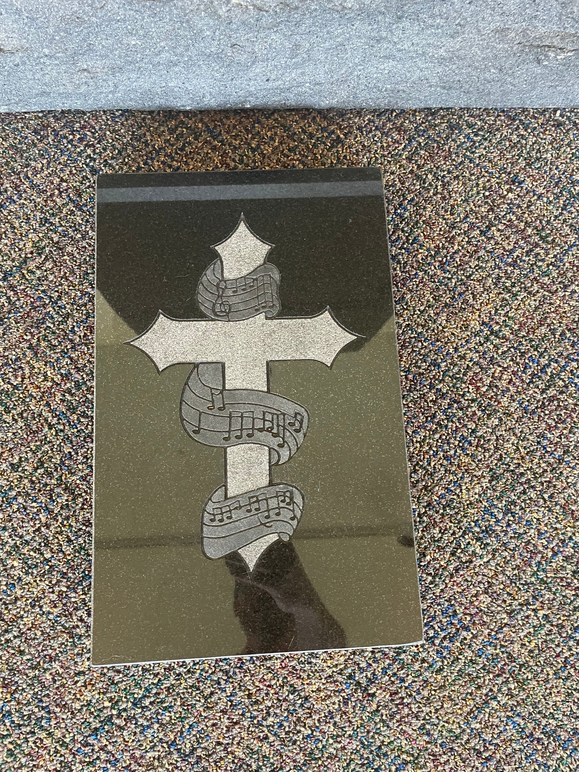 Dark granite headstone with a silver cross intertwined with a serpent. The headstone rests on a speckled carpet.