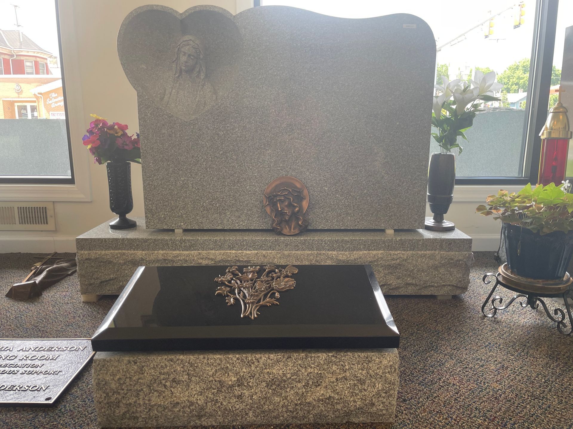 A granite tombstone with an angel, flowers, and a black headstone, inside a building with a window.