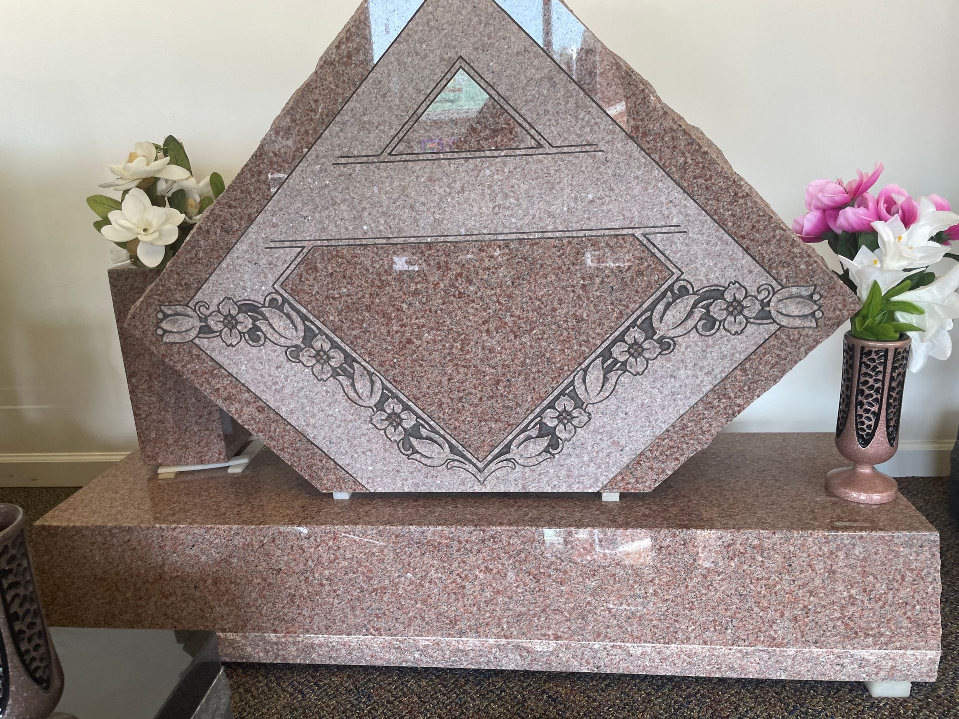 A large, pink granite tombstone with a diamond-shaped center. Floral arrangements flank it.