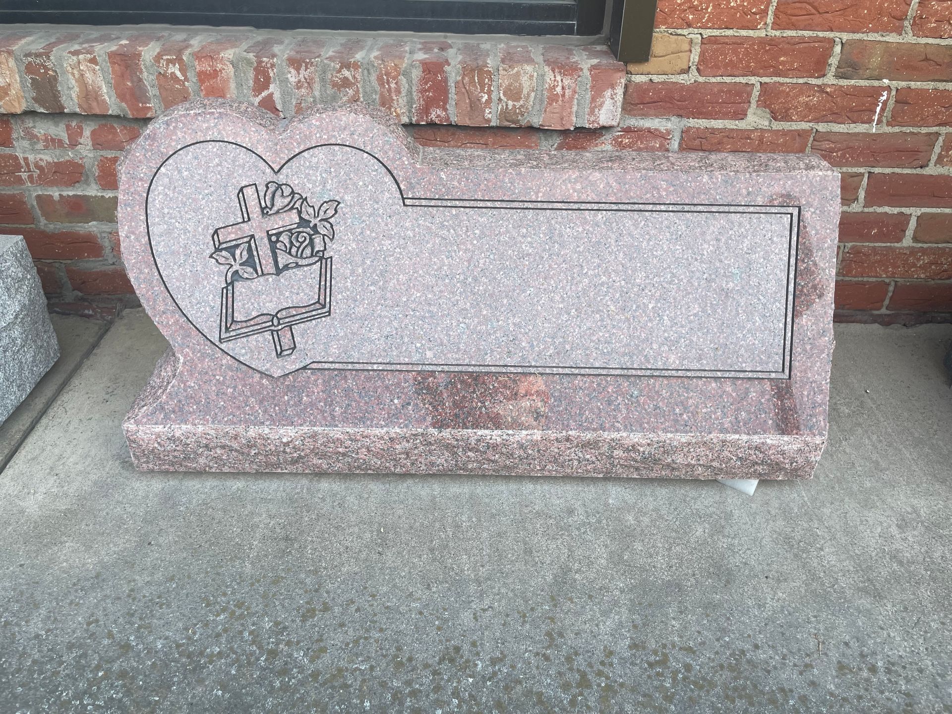 Pink granite headstone with heart-shaped top, a carved design, and a blank rectangular area for text.  It rests on a concrete surface against a brick wall.