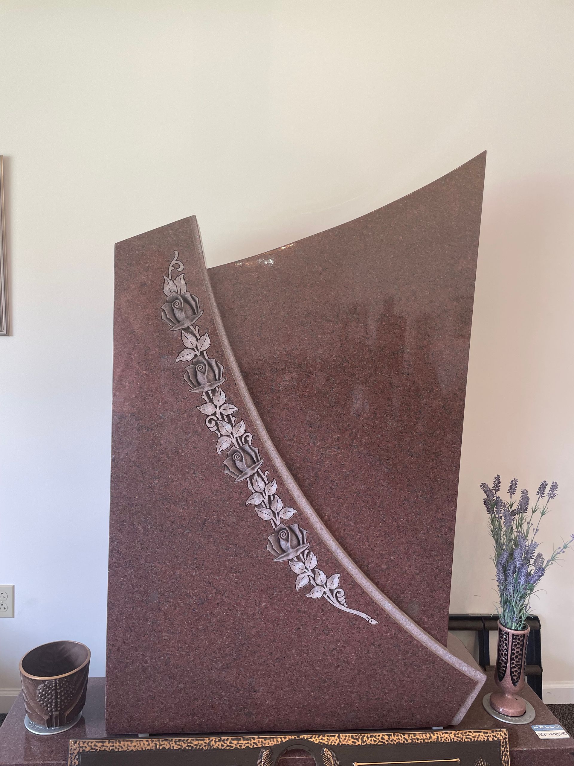 A reddish-brown granite memorial with an arched, curving shape. It features an etched floral design and stands indoors near a vase of flowers.