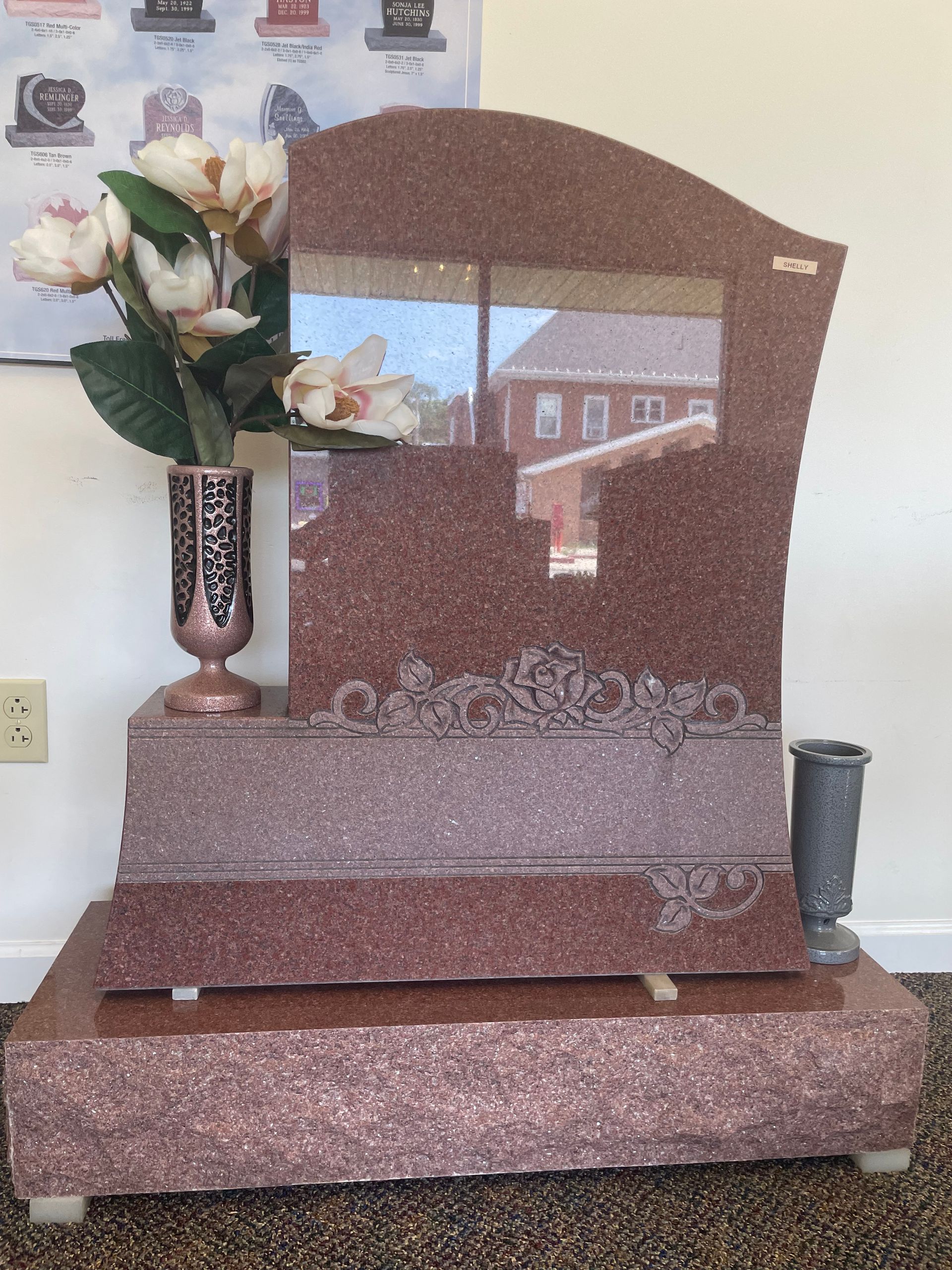 A red granite headstone with a vase of flowers and a small vase. The stone is reflective and has decorative etchings.