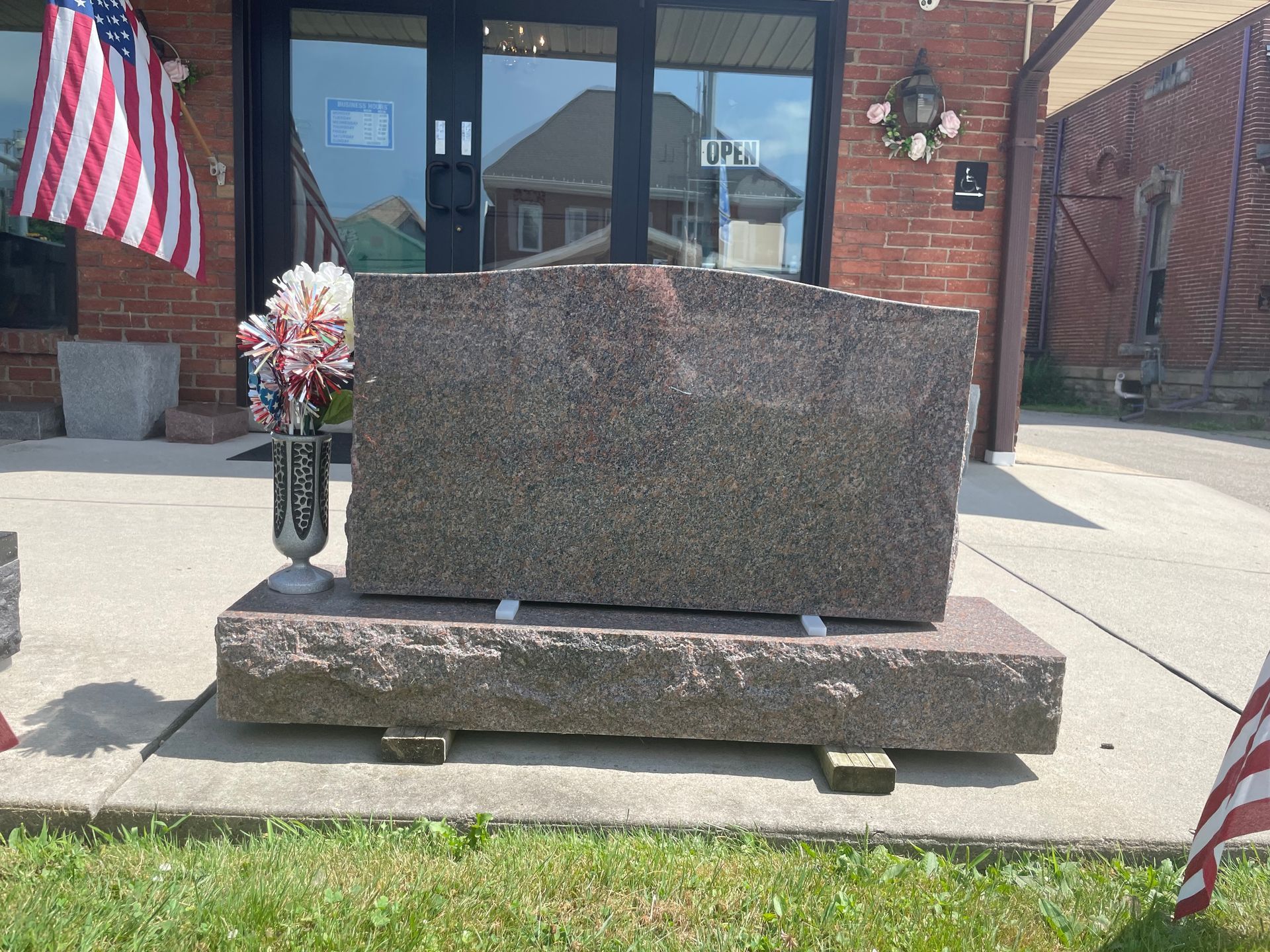 A large granite tombstone rests on a base, displayed outside a brick building with an American flag.