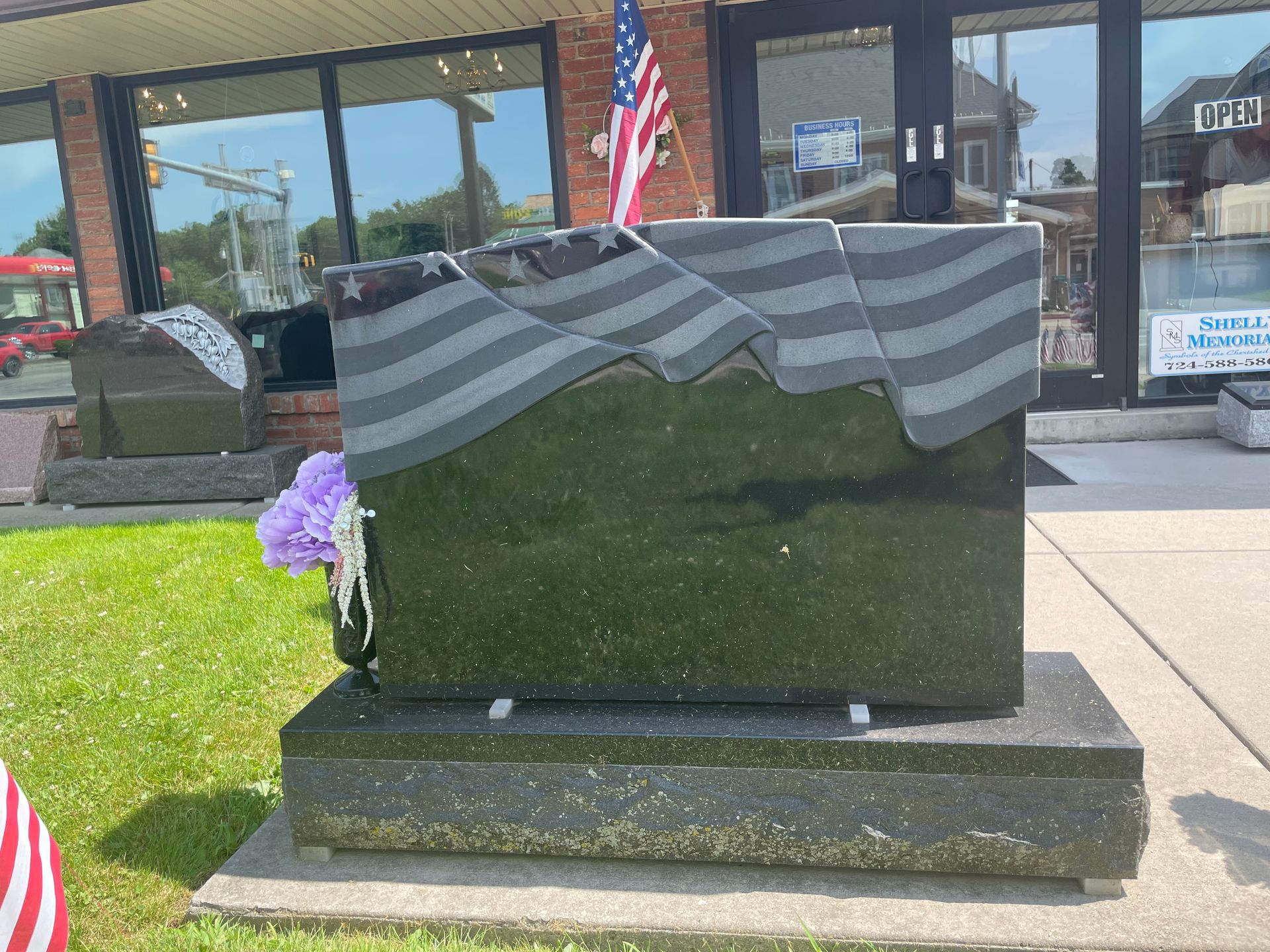 A black granite memorial with a waving American flag design; a small bouquet of purple flowers rests at the side.