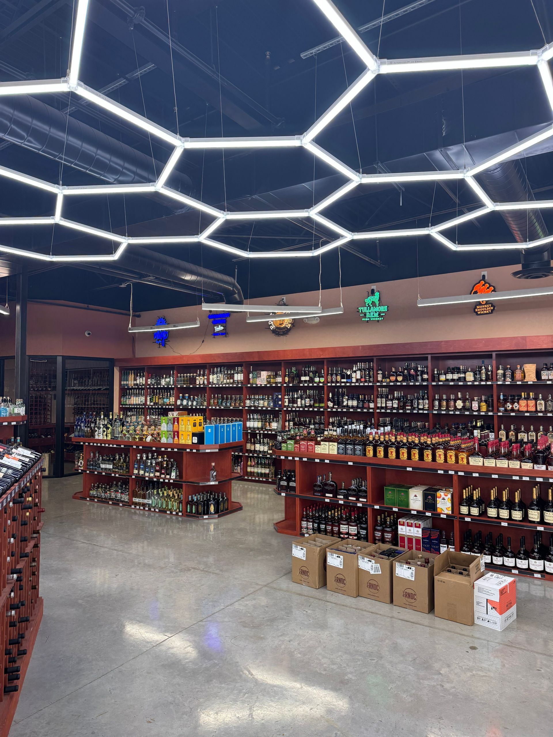 Liquor store interior: shelves stocked with bottles, overhead honeycomb lights, and a concrete floor.