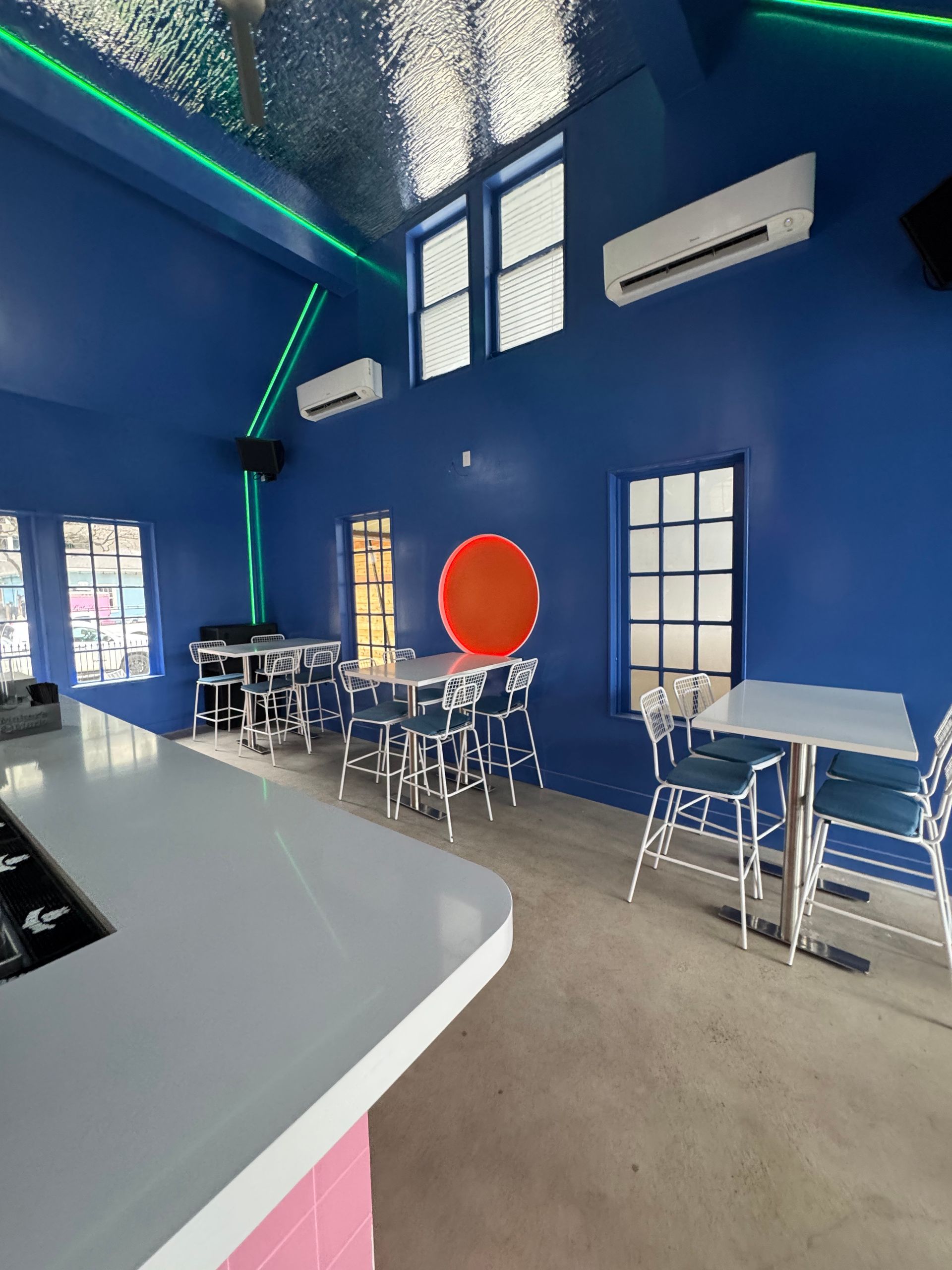 Blue-walled interior with white bar, tables, chairs. Orange circle art. Fluorescent green lighting.