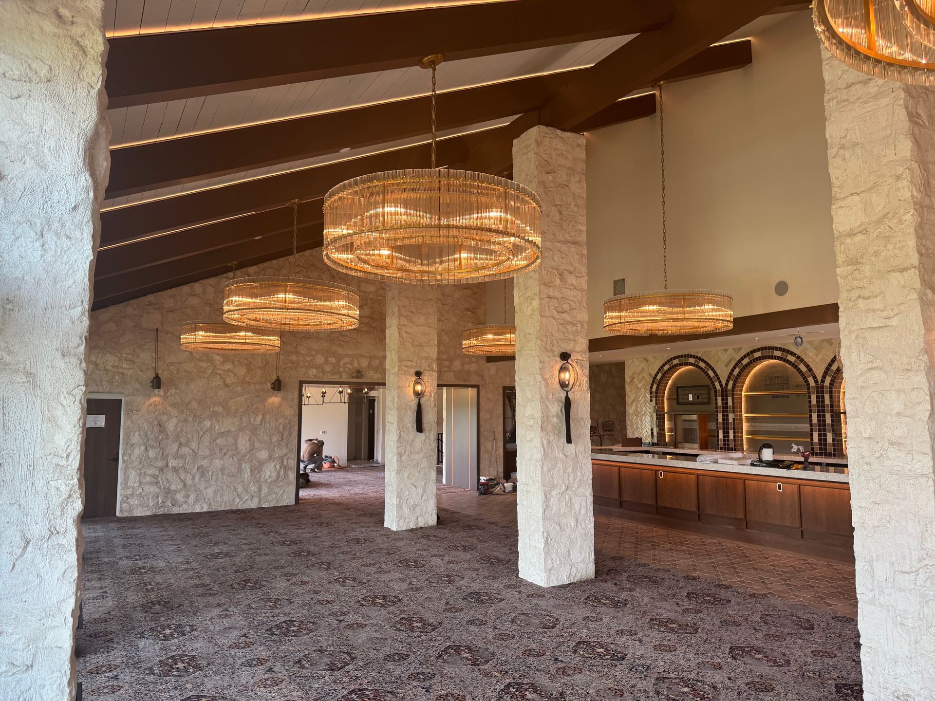 A large event space with stone columns, brown beams, and decorative lighting over a patterned carpet.