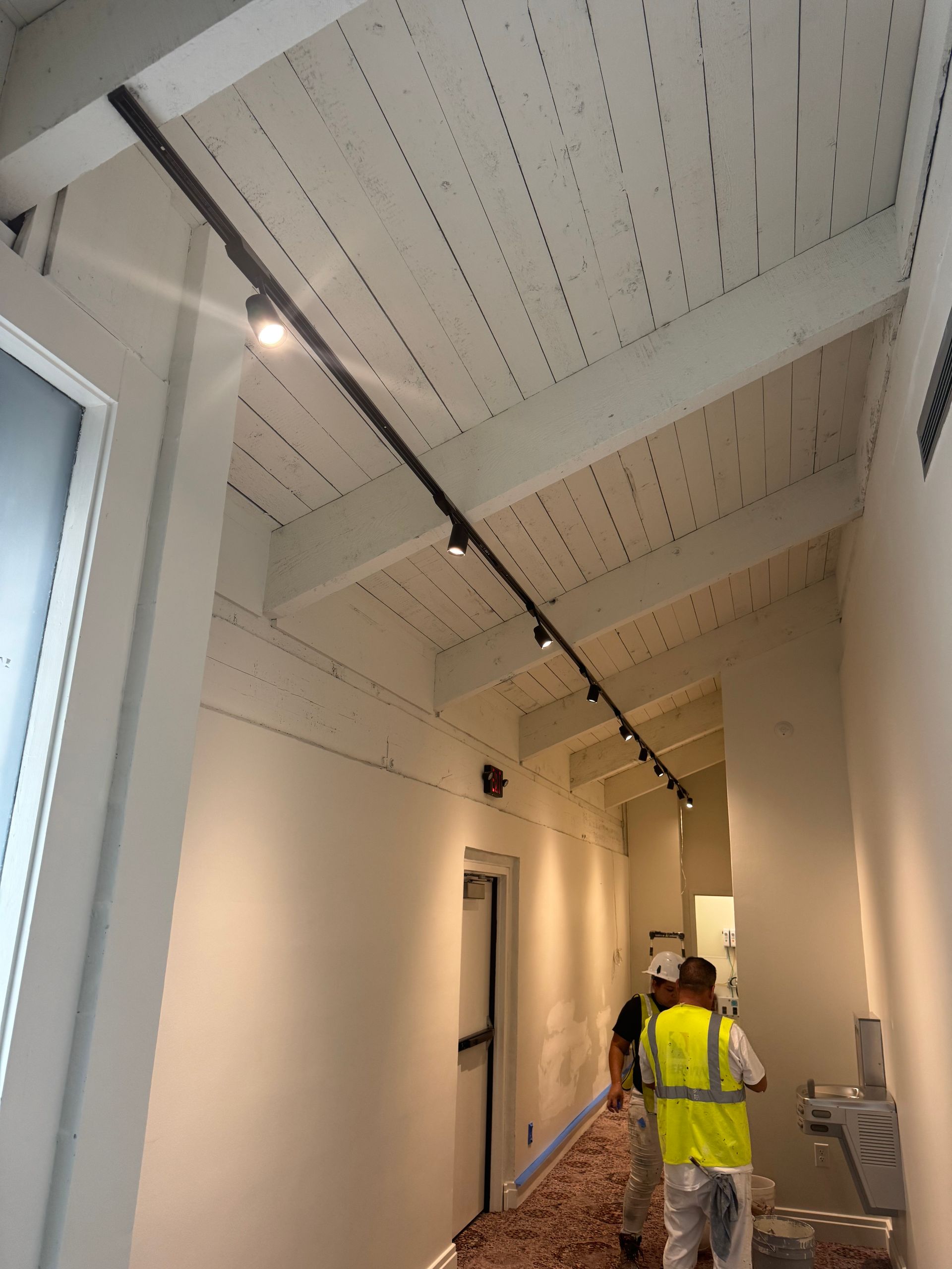 Narrow hallway with white walls and ceiling, track lighting. Two workers in neon vests.