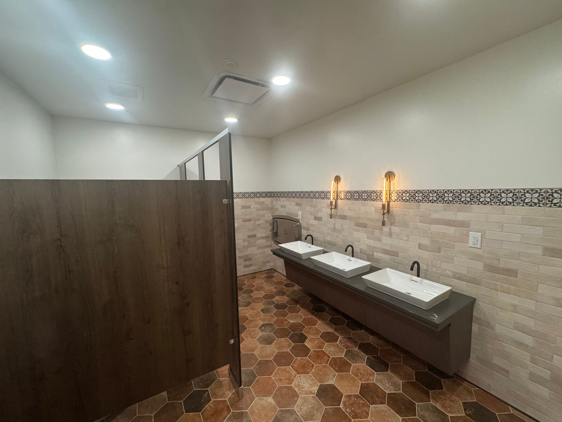 Public restroom with three sinks, wood stalls, patterned tile floor, and decorative wall.