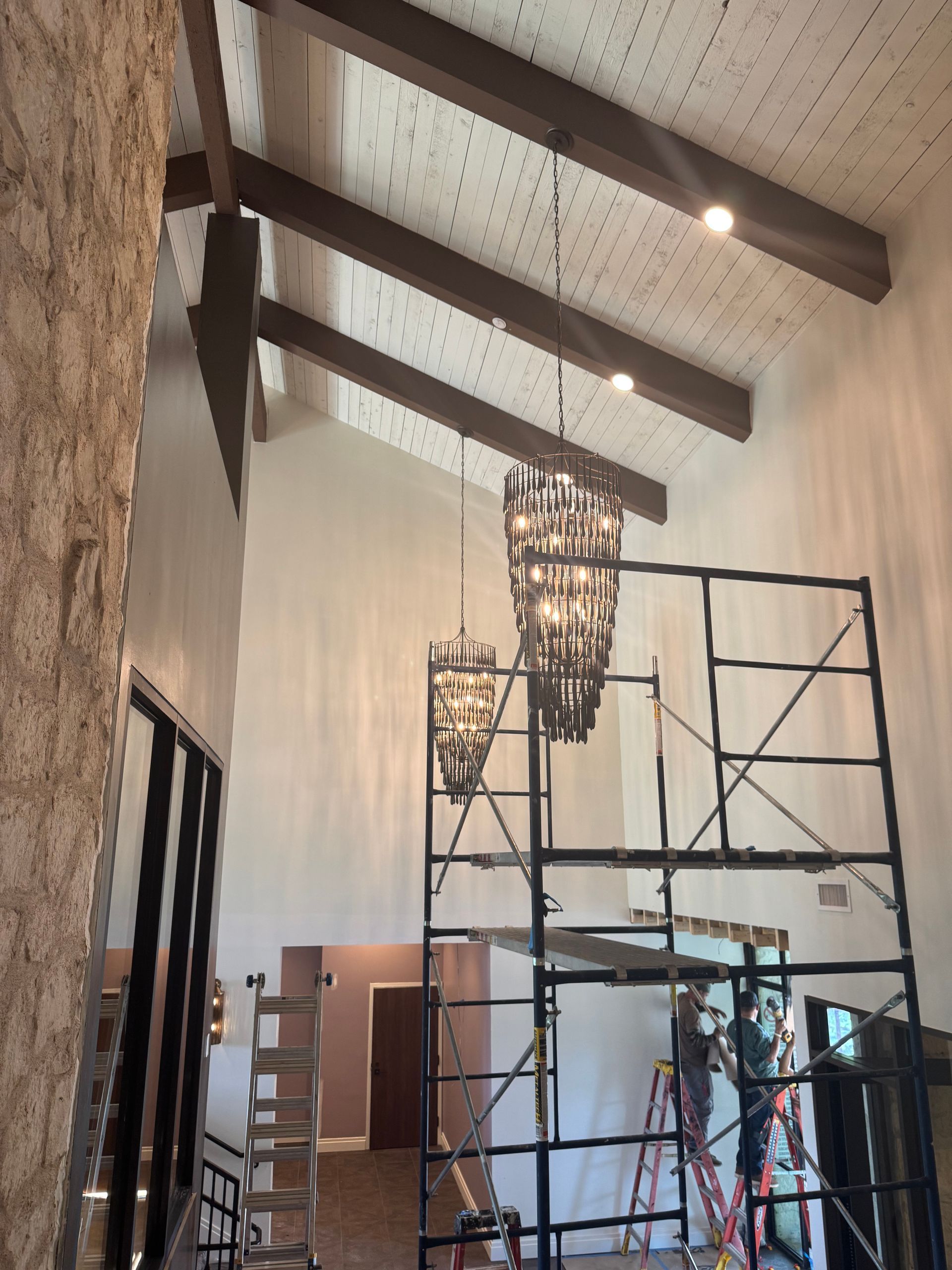 High-ceilinged room with dark wood beams and two chandeliers. Scaffolding supports workers, one on a ladder.