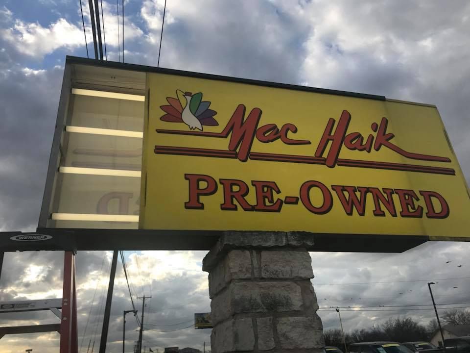 Sign for Mac Haik Pre-Owned, yellow with red text, a peacock logo, and a brick base under a cloudy sky.