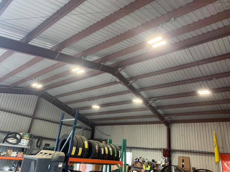 Inside a metal building, lights illuminate a cluttered workspace with tires and shelving.