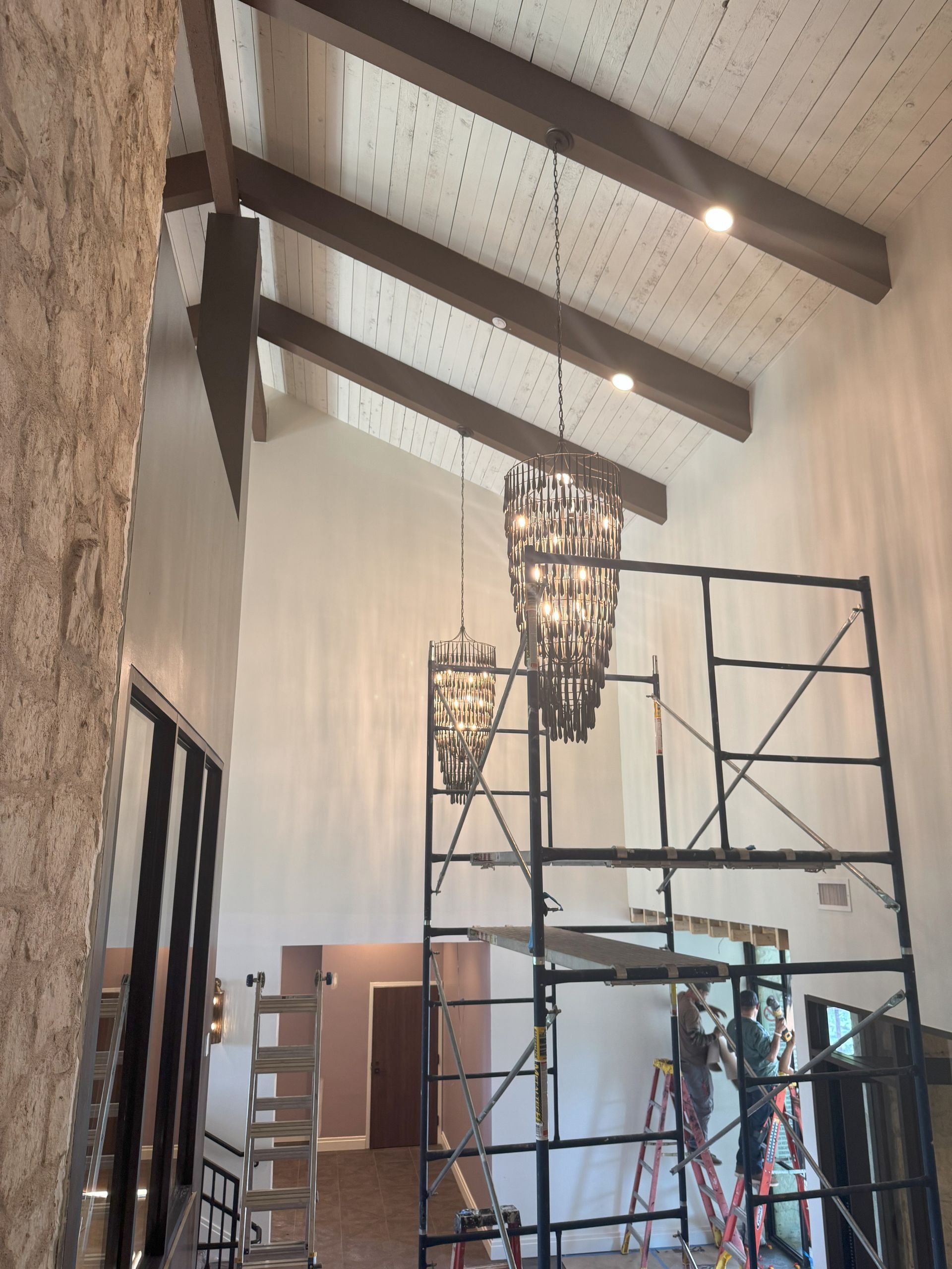 High-ceilinged room with scaffolding, two chandeliers, exposed beams, and workers.