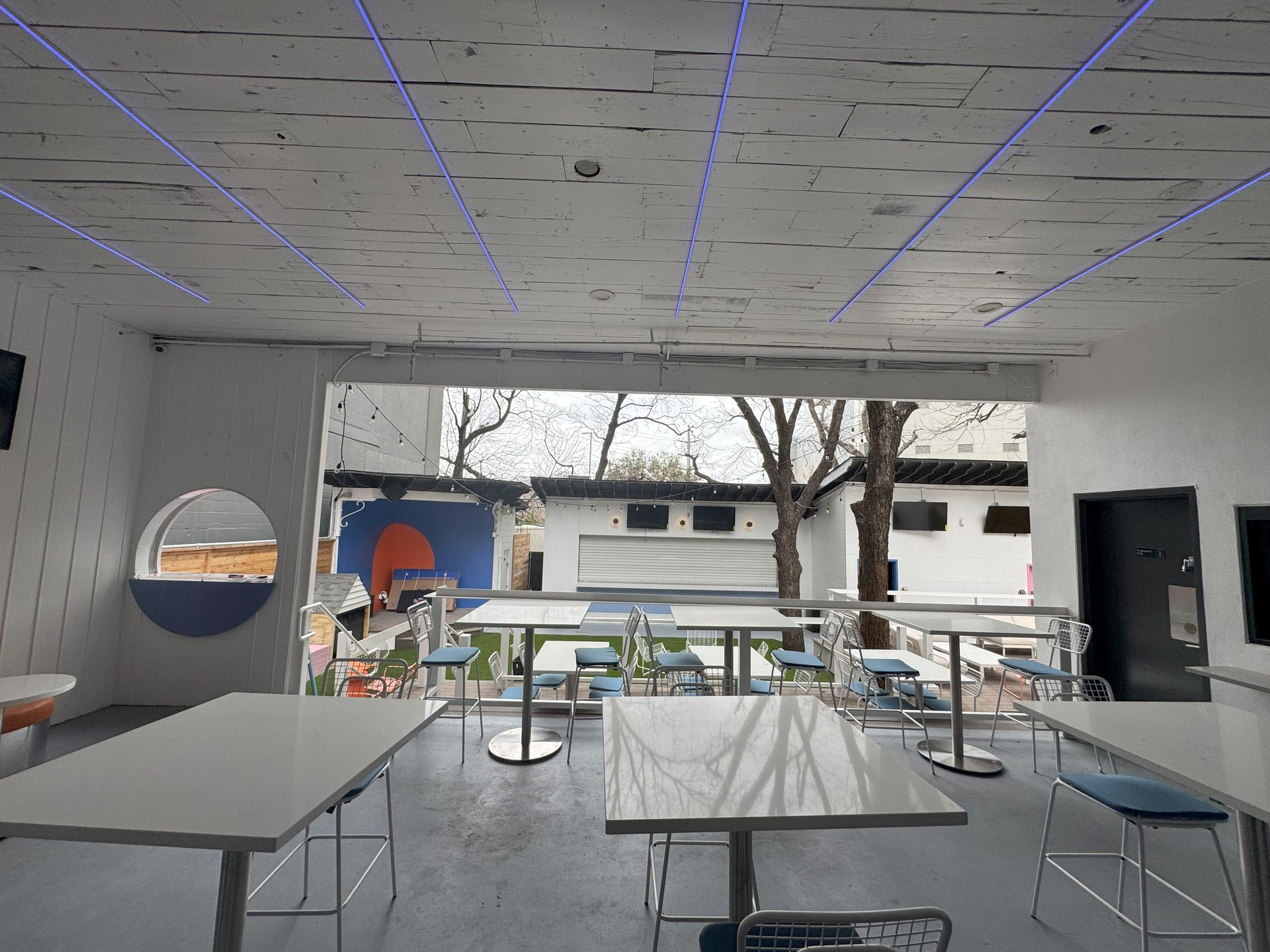 Interior dining area with tables, looking out to an outdoor seating area. The ceiling has blue lights.