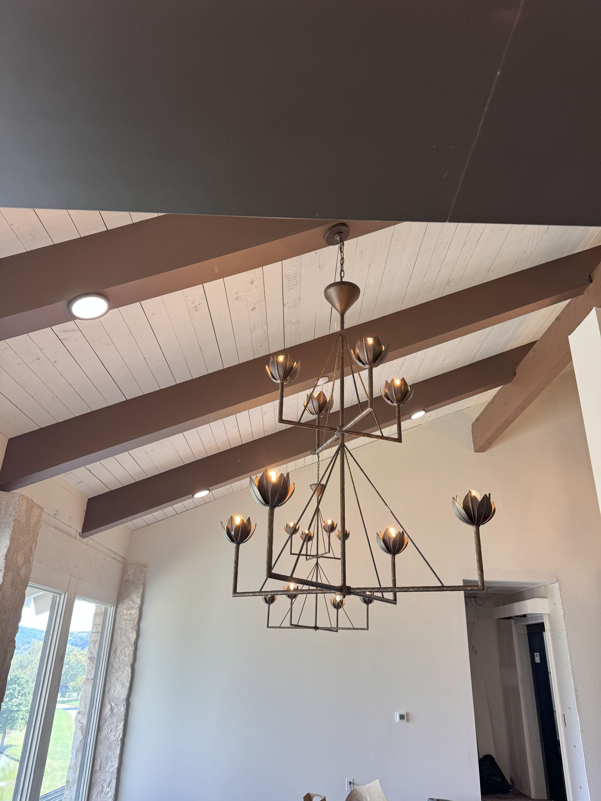 Chandelier hangs from a brown beamed ceiling in a room with a window and doorway.