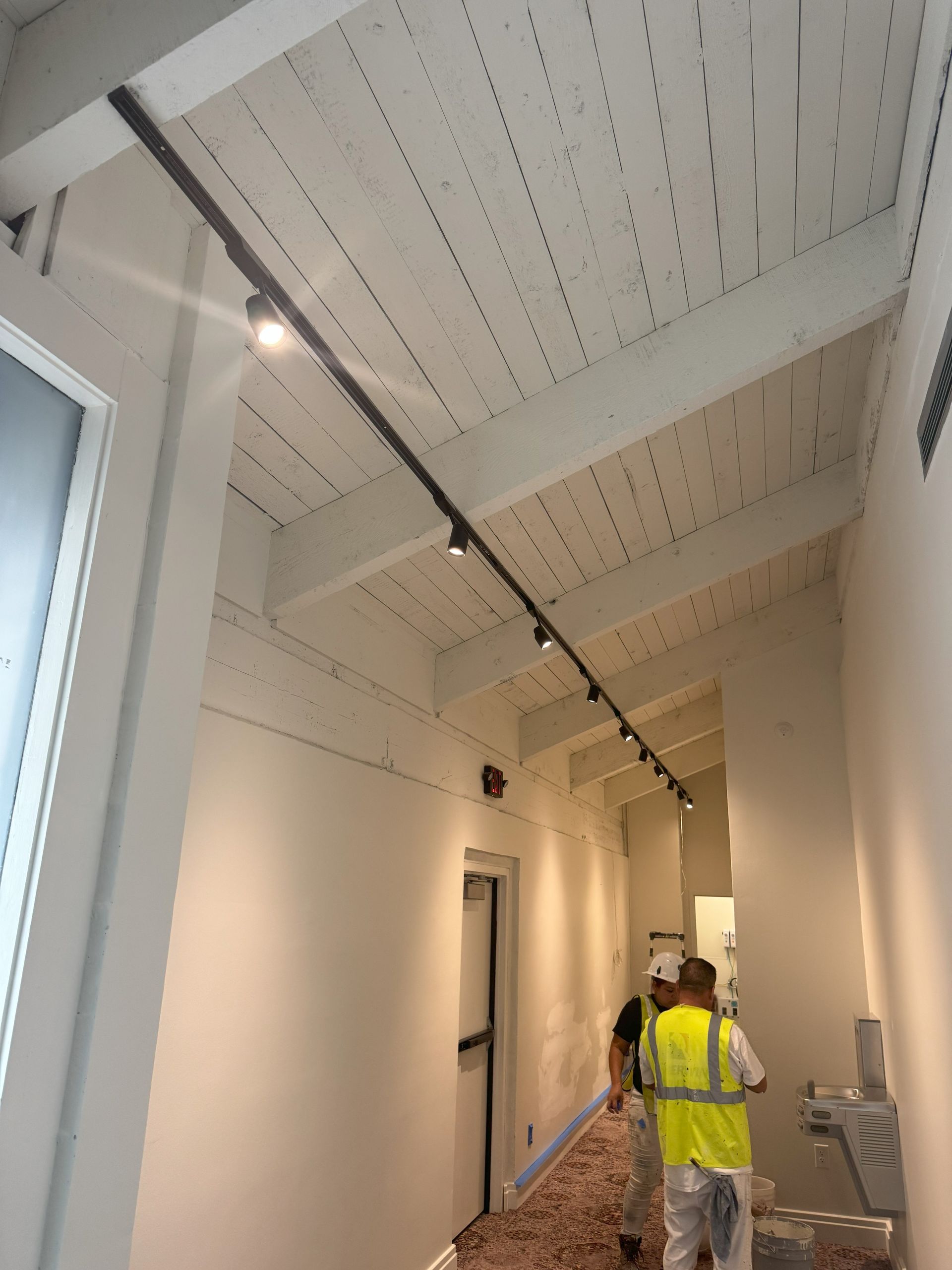 Hallway with track lighting on sloped white ceiling; two workers wearing yellow vests.