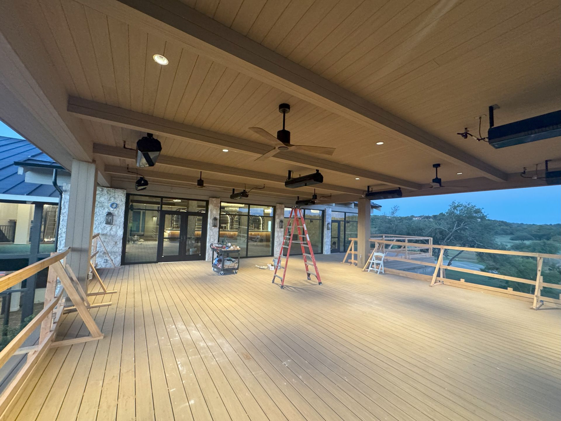 A covered outdoor deck with overhead lighting, fans, and a distant view. A ladder and railings are present.