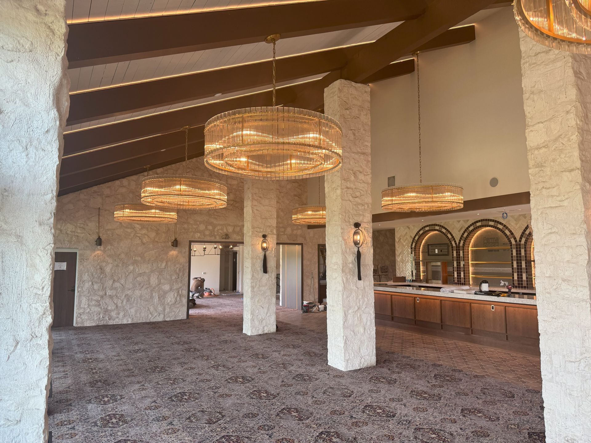 Spacious room with brown beams, tan pillars, chandeliers, bar area. Brown carpeted floor.