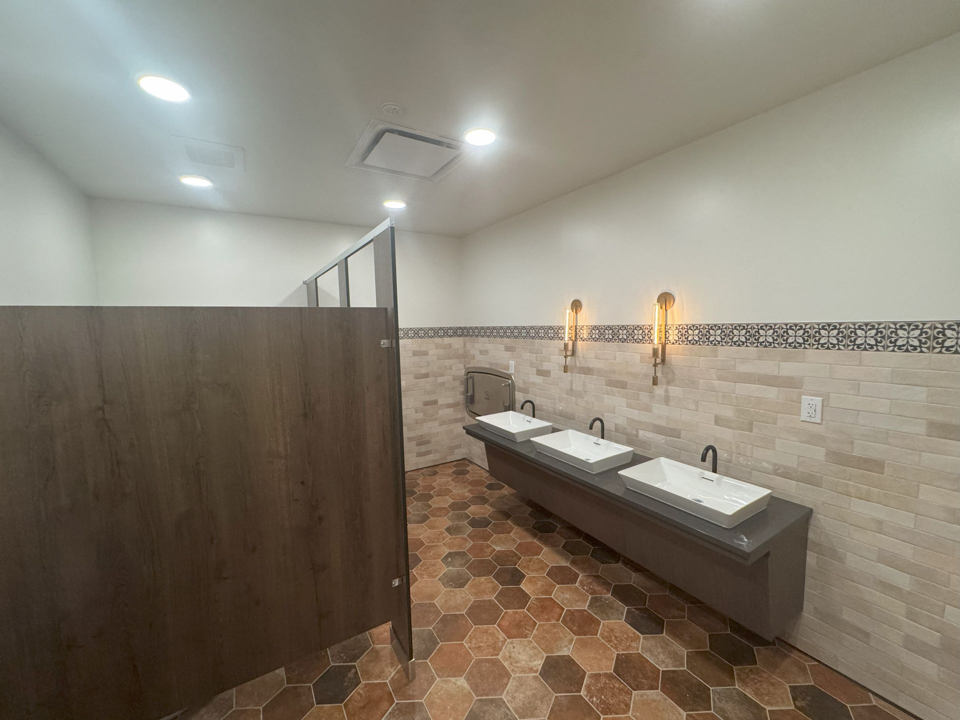 Public restroom with three sinks and stalls, tile floor, cream walls, wood stall doors.