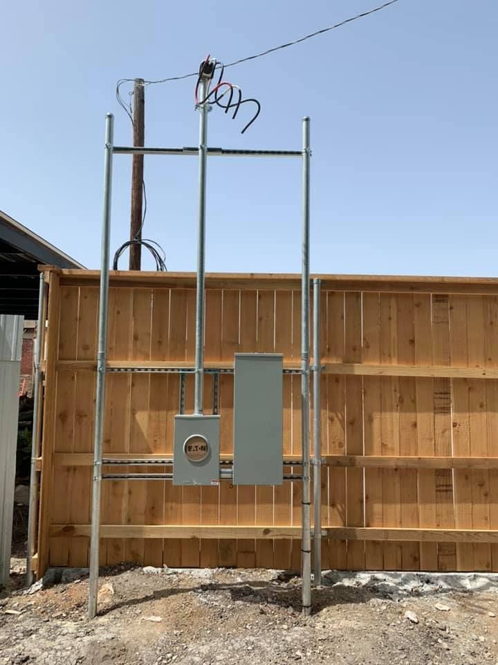 Electrical utility installation with meter box, conduit, and wires, mounted on metal supports next to a wooden fence.