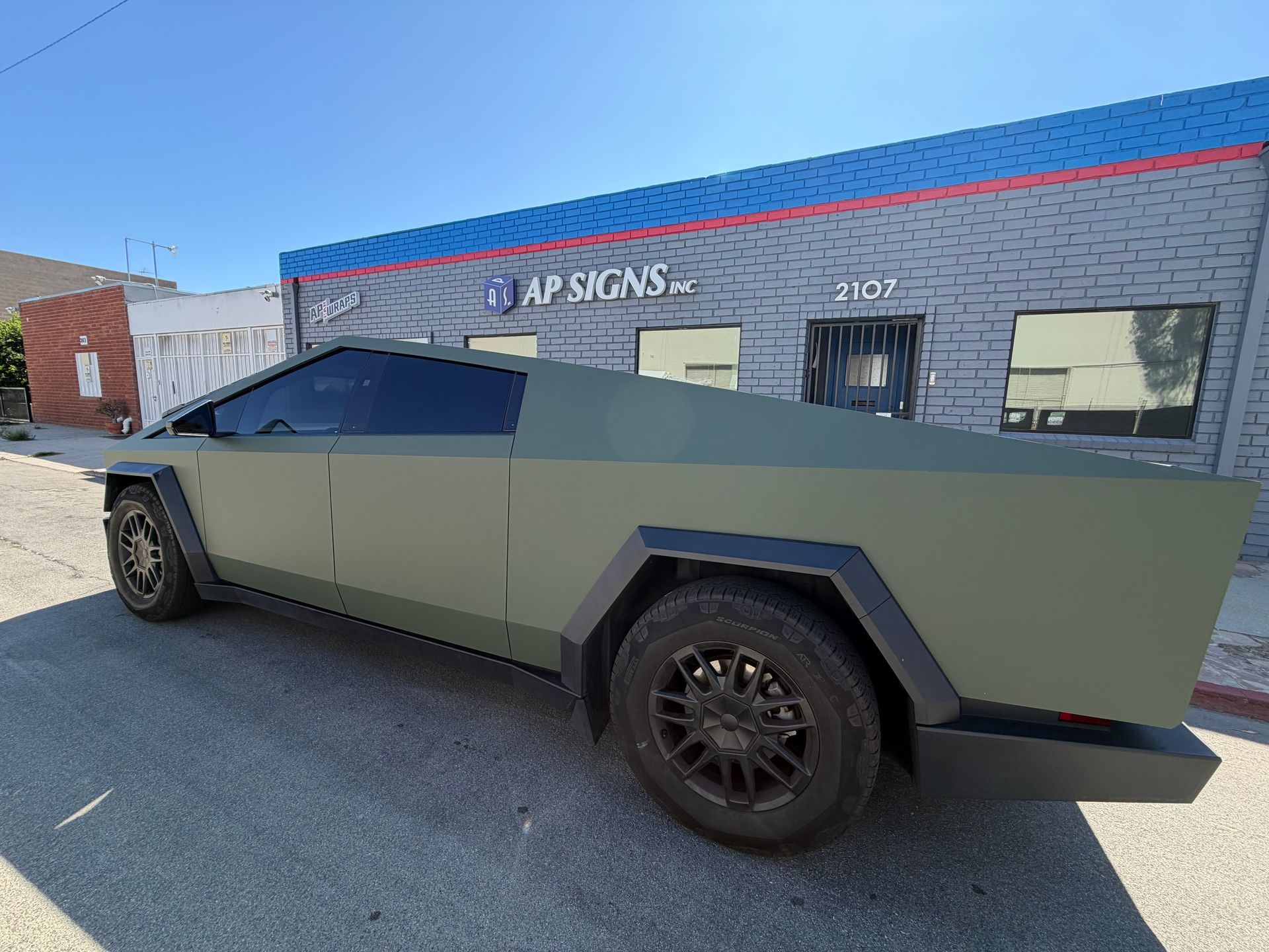 Matte green angular pickup truck parked outside a storefront on a sunny street