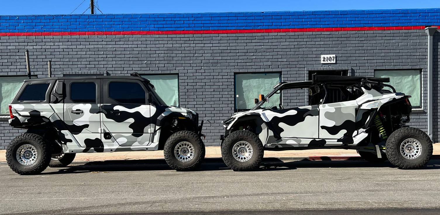 Two camouflaged SUVs parked side by side against a gray brick wall