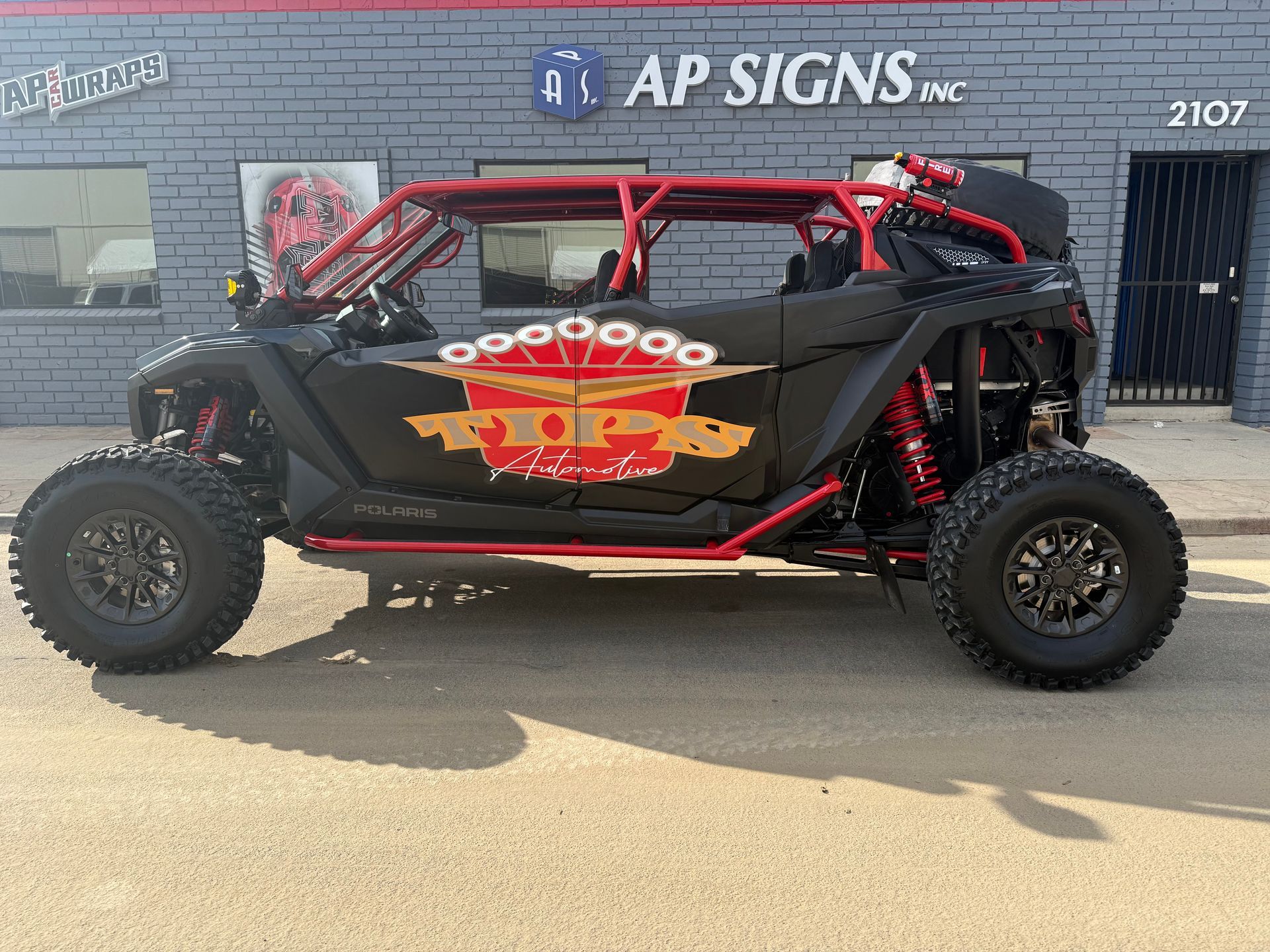 Black off-road buggy with red roll cage parked outside AP Signs storefront.