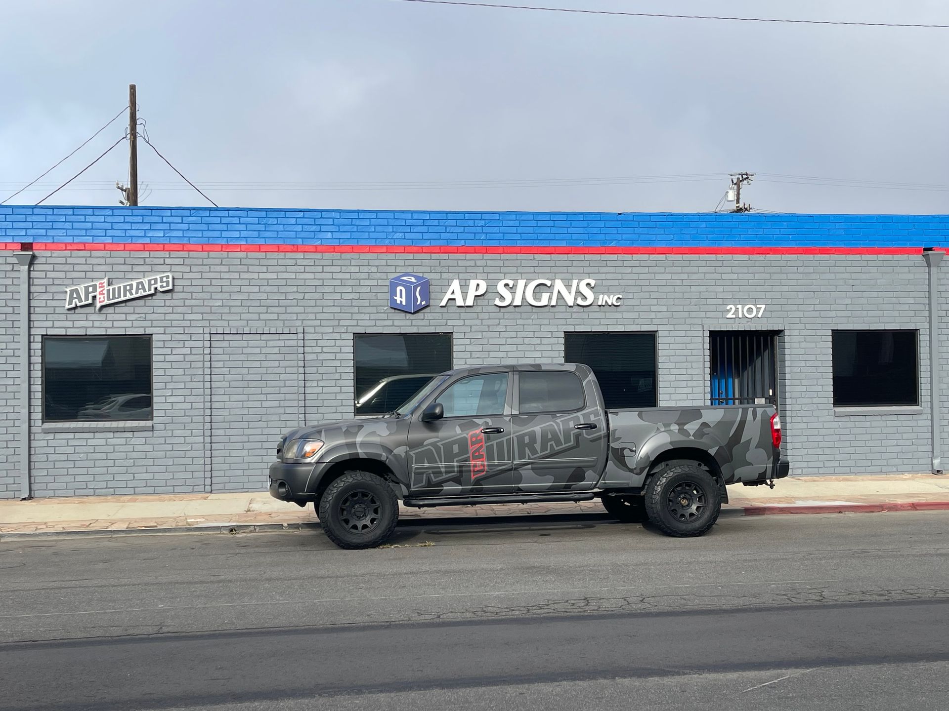Damaged gray pickup truck parked outside AP Signs storefront with red and blue trim