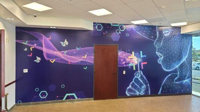 Wall mural in an office space with a figure blowing colorful digital shapes. Dark blue background, geometric patterns, and butterflies.