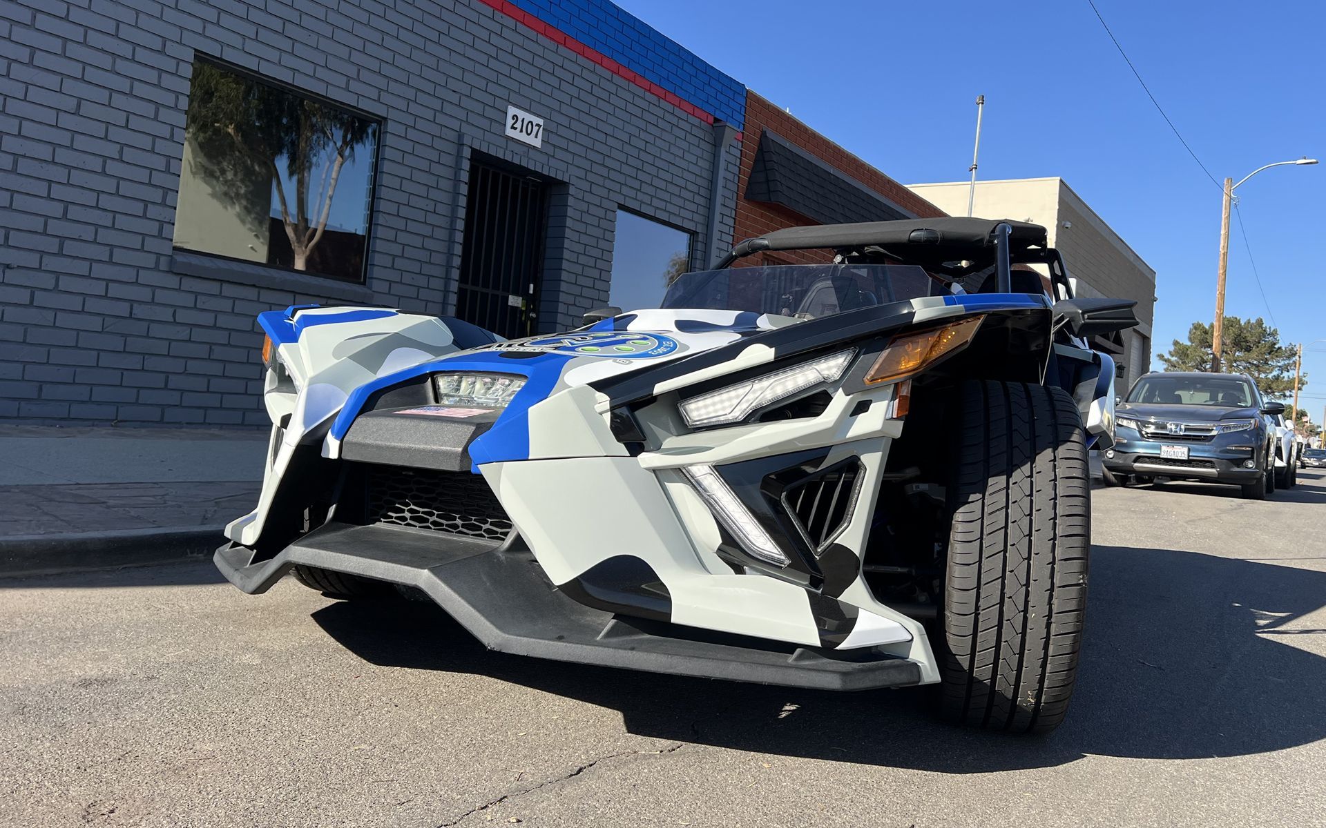 A white and blue slingshot is parked in front of a brick building.