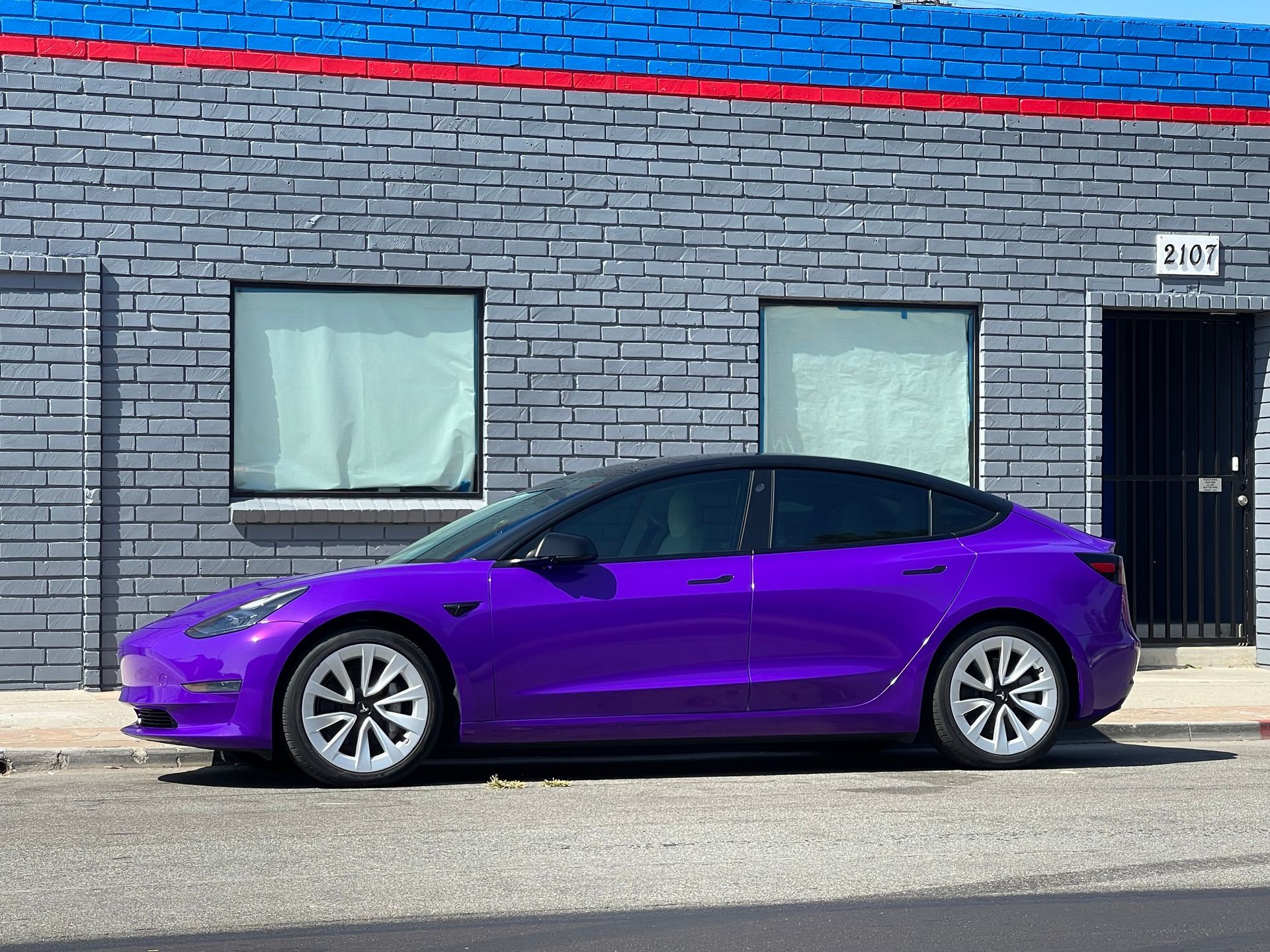 A purple tesla model 3 is parked in front of a brick building.