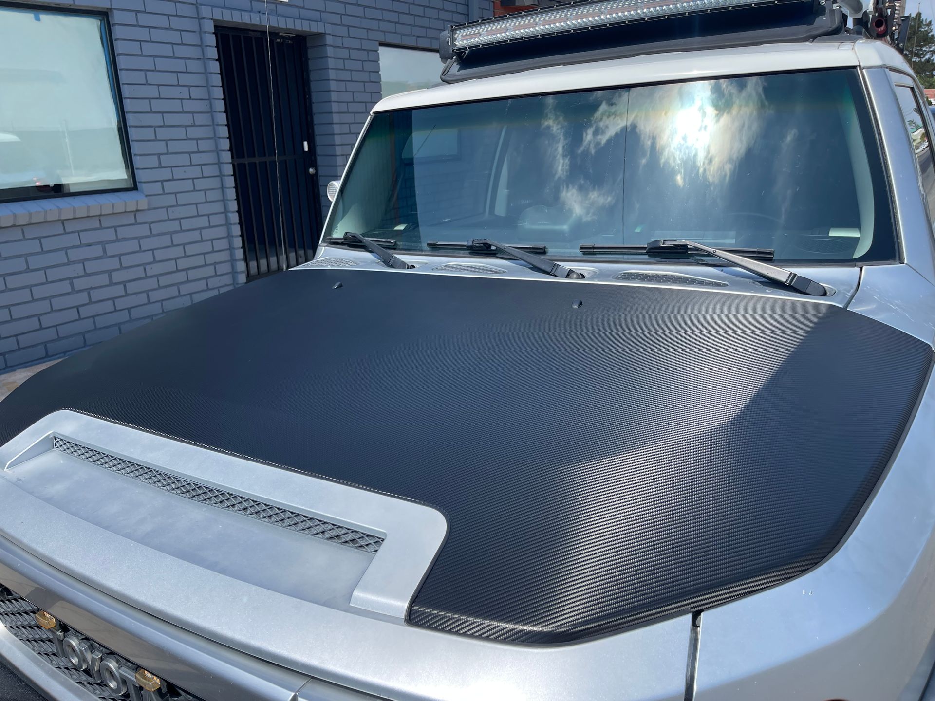 A silver truck with a black hood is parked in front of a brick building.