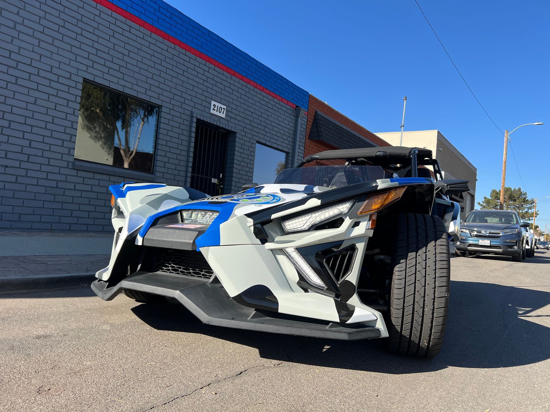 A white and blue slingshot is parked in front of a brick building.