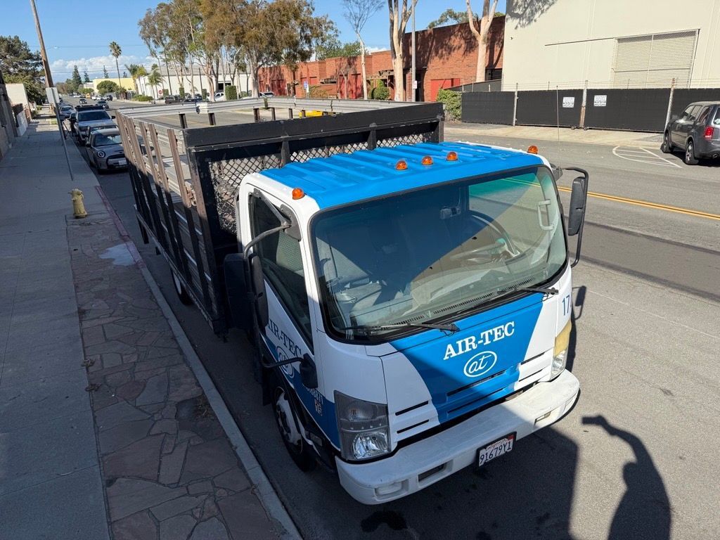 White and blue Air-Tec truck parked on a street, bed filled with debris. Sunny day, cars parked nearby.