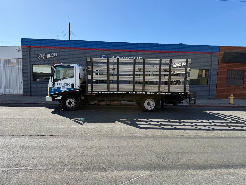 White truck with wooden rails parked on street in front of a brick building under a blue sky.