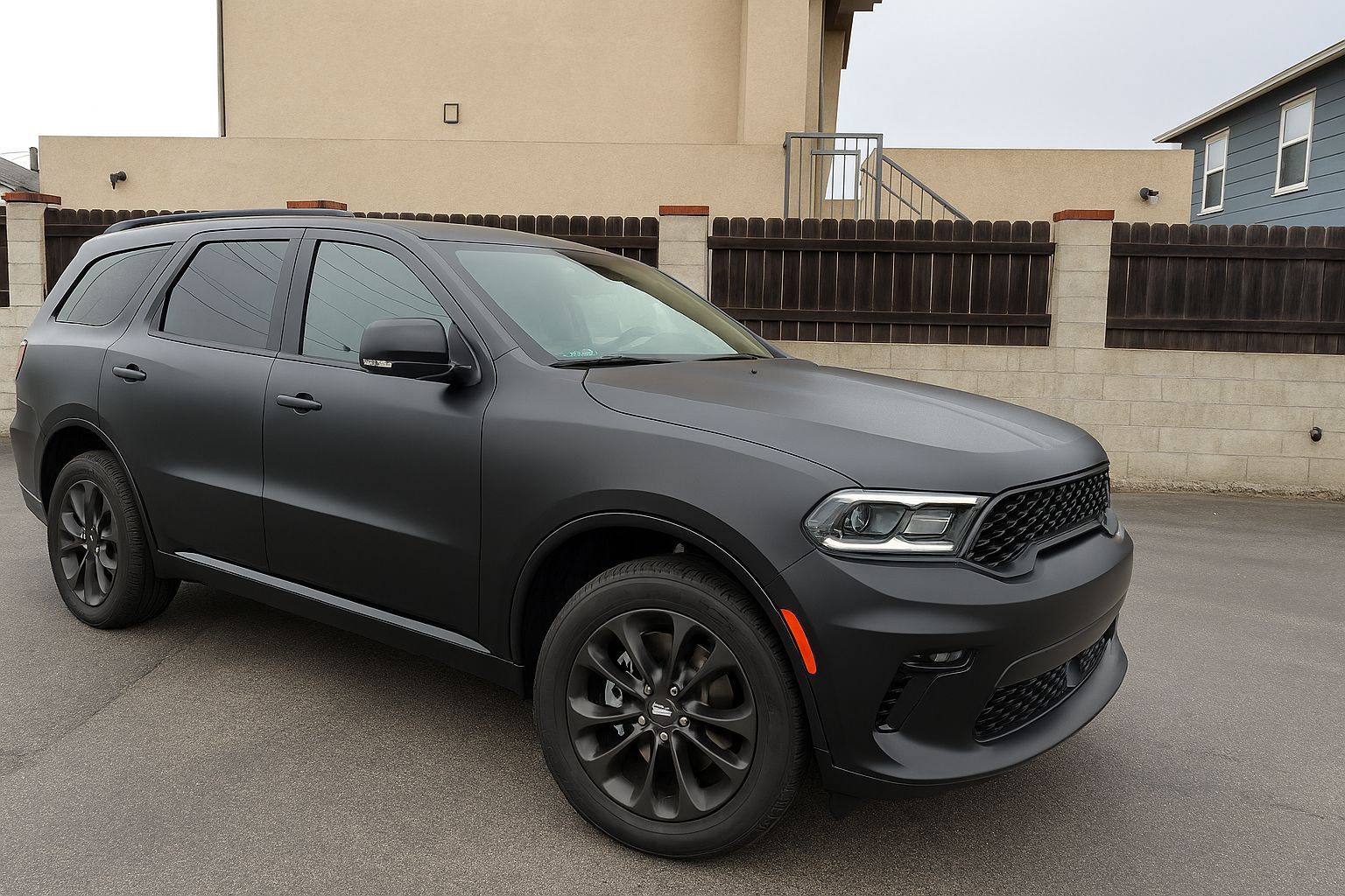 Black Dodge Durango SUV parked on asphalt. Black wheels, tinted windows. Beige building and fence in the background.