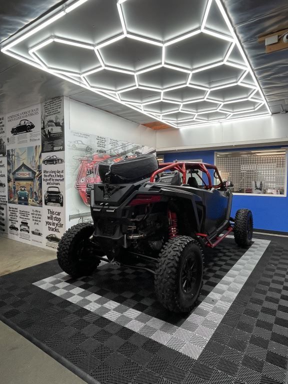 Off-road vehicle on black and gray checkered floor, under honeycomb lights in a garage-like space.