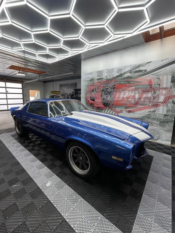 Blue and white classic Camaro with racing stripes in a well-lit garage, with a mural on the wall.