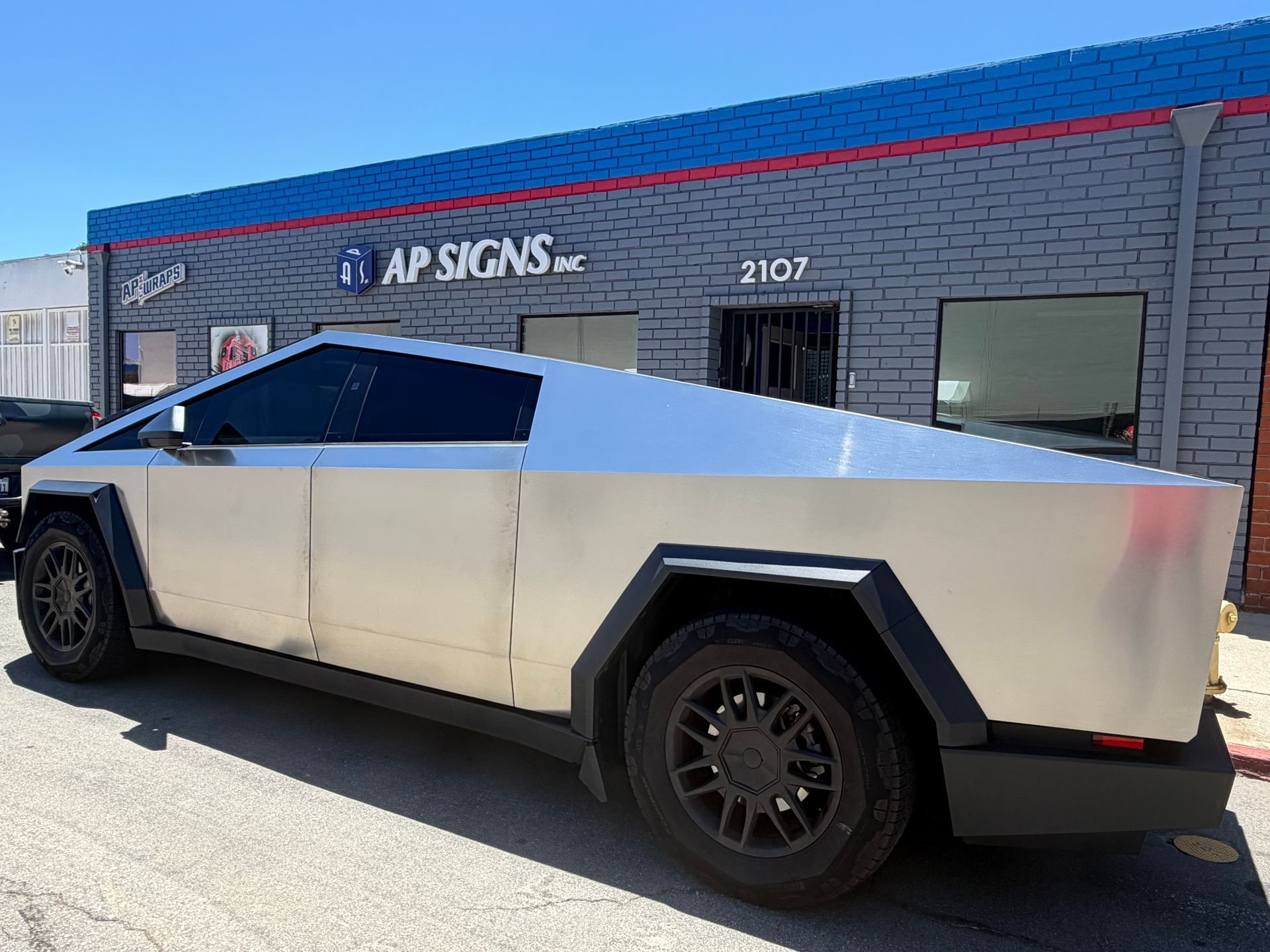 Silver Tesla Cybertruck parked outside AP Signs storefront in bright sunlight