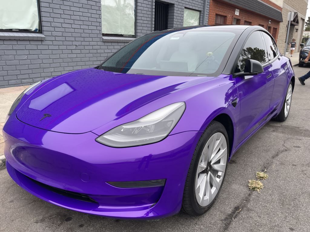 Purple Tesla parked on a street beside a gray brick building.