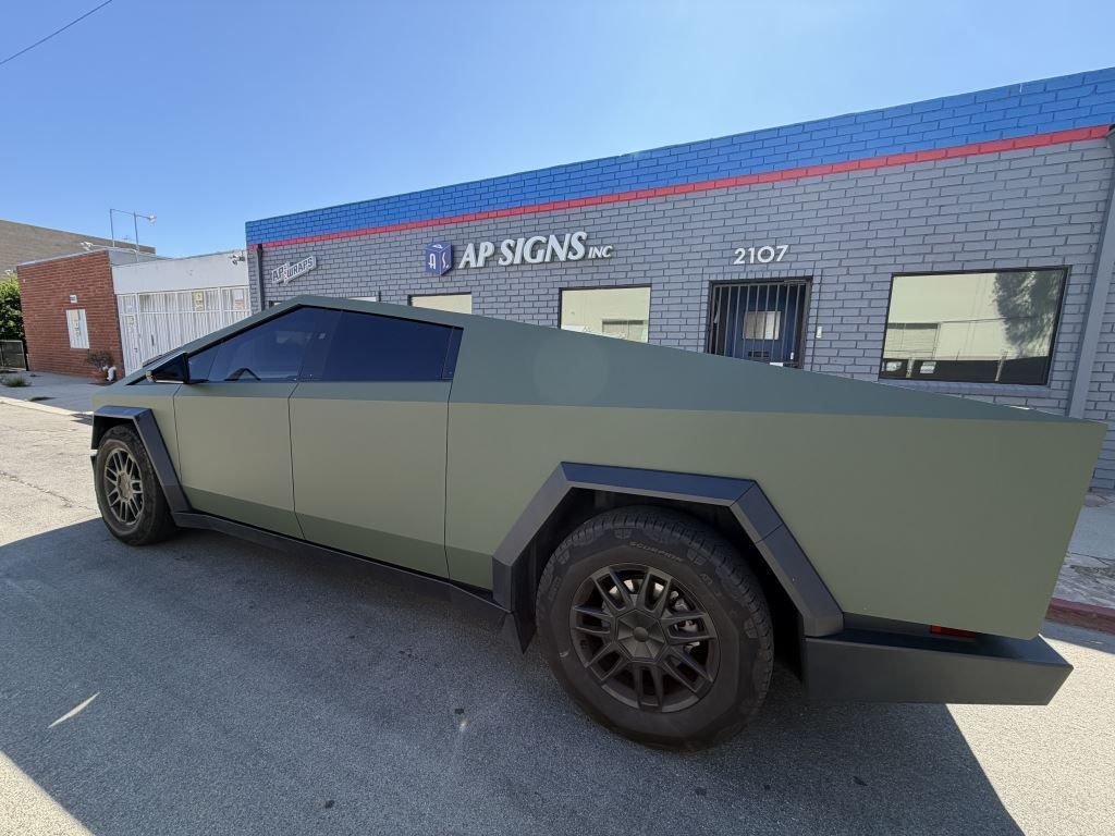 Matte olive-gray Cybertruck parked outside a building with AP Safety signs