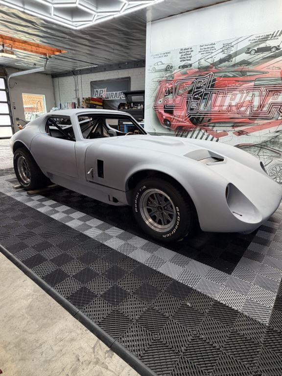 Silver classic race car displayed indoors on a checkered floor, with a red mural behind it