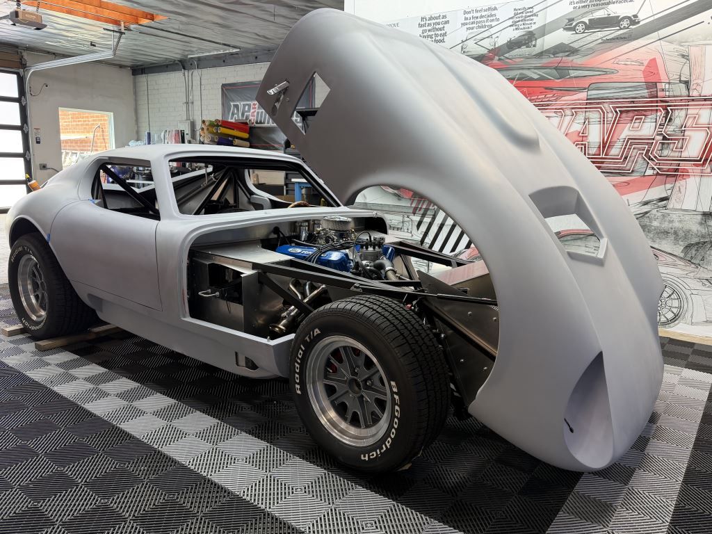 Classic car in a workshop with its front body shell lifted, exposing the engine and chassis.