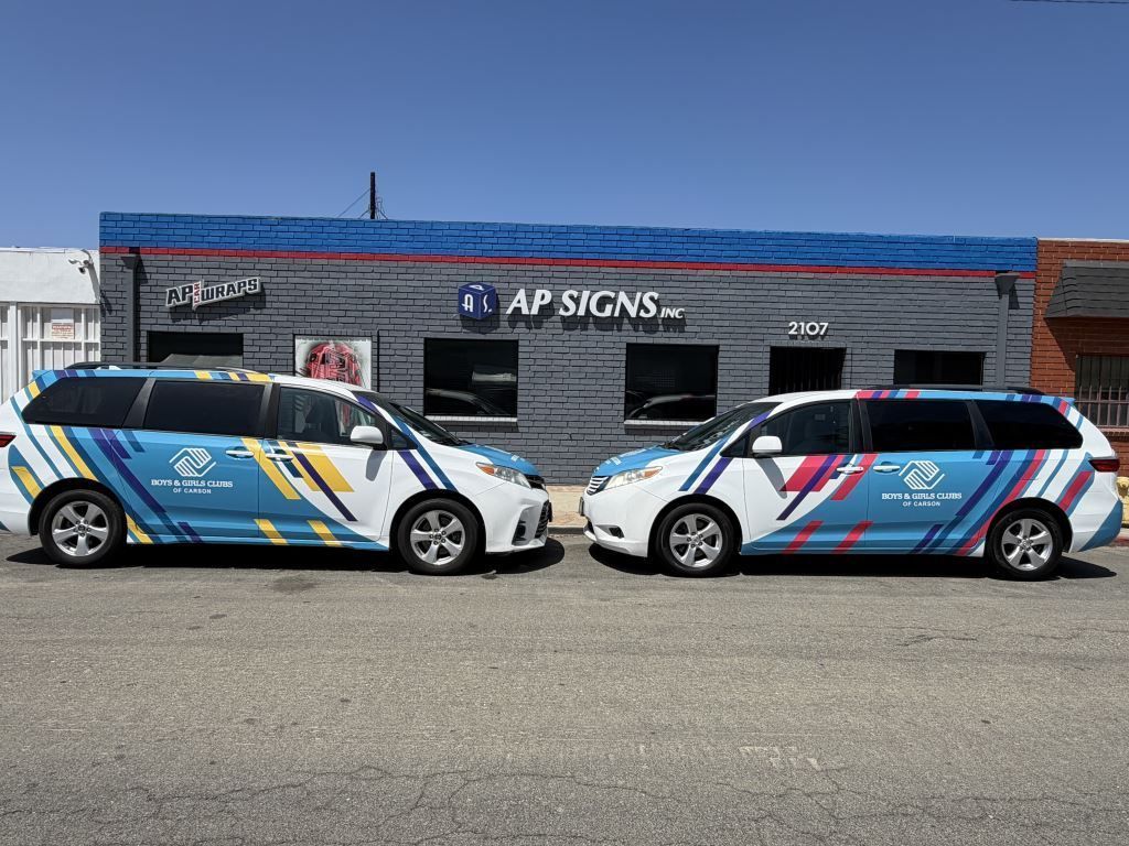 Two branded white hatchbacks parked outside AP Signs storefront with blue and pink graphic wraps