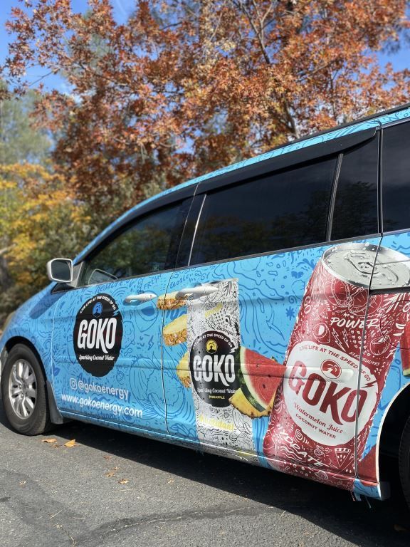 Blue minivan wrapped in colorful OKO soda ads parked outdoors by autumn trees